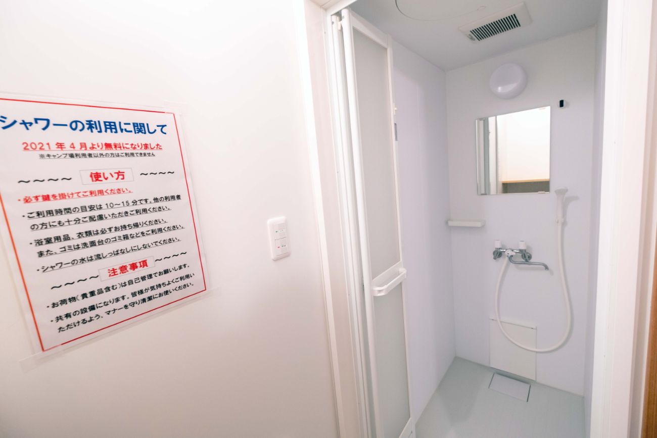 シャワールームの利用案内が書かれた掲示板と白い壁に囲まれ、鏡とシャワーが設置されているシャワールームを写した写真