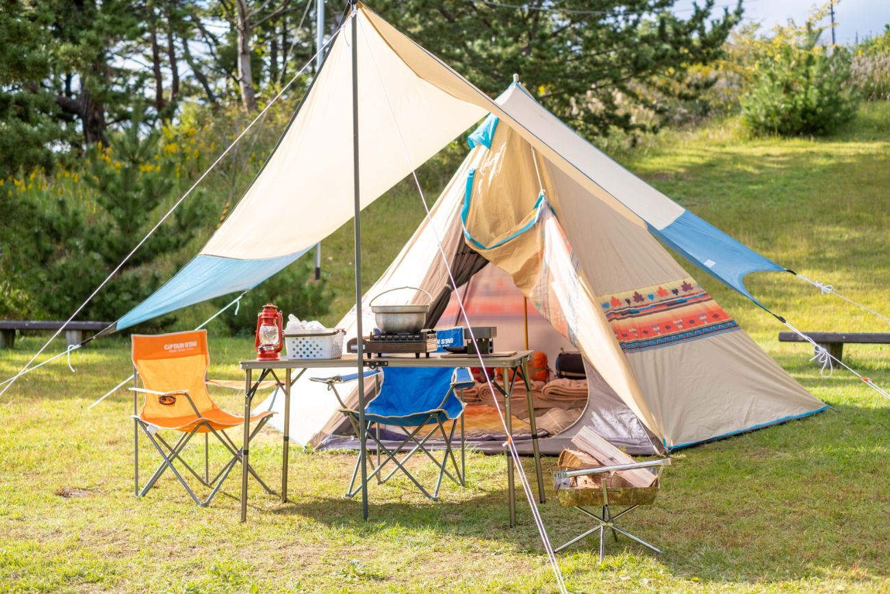 草地に立てられたベージュと青のテントの前に、オレンジ色と青色の折りたたみ椅子が向かい合って置かれ、その間にあるテーブルの上にガスコンロと鍋、赤いランタン、白いカゴが並べられている屋外キャンプ場の写真