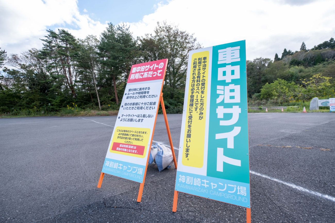 舗装された広い駐車場の一角に、看板が2枚並び、キャンプ場の車中泊サイトの利用に関する案内が記載されている「神割崎キャンプ場」の看板の写真