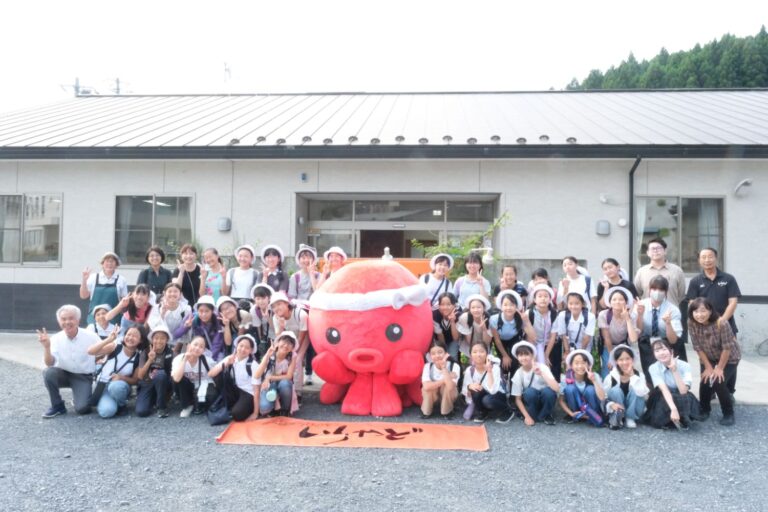 白い建物の前に大きな赤いタコのキャラクターオクトパス君が座っており、その周りに多くの人々が並んでいる集合写真