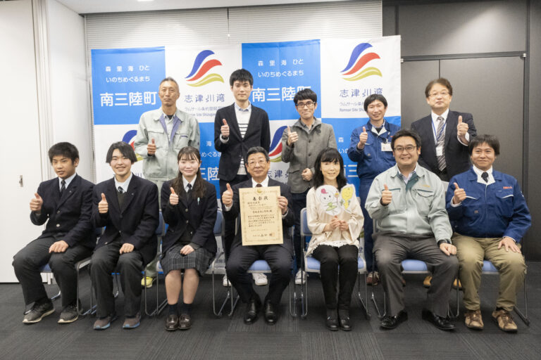 南三陸町のバックパネルの前で、12人の人々が2列に並び、前列の中央に座る佐藤町長が証書を持ち、ほとんどの人が親指を立ててポーズをとっている様子を写した写真