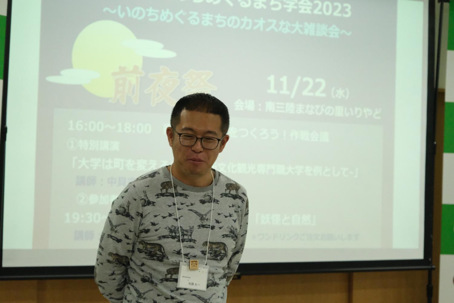 男性がスクリーンの前に立っており、スクリーンには「いのちめぐるまちのカオスな大討論会」などの文字が表示されている写真