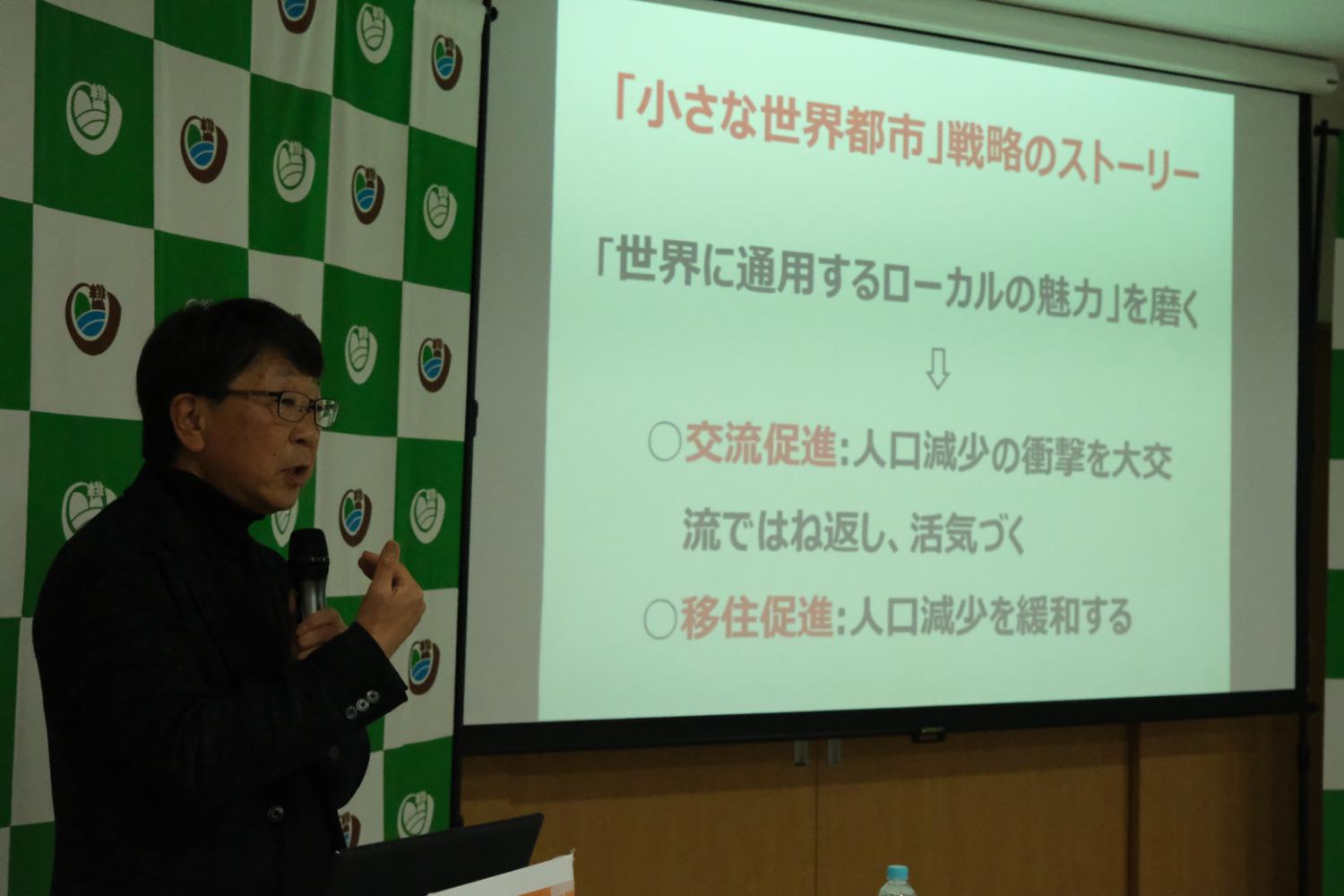 講演会場で中見宗治さんがマイクを持って話しており、参加者たちが話に耳を傾けている様子の写真