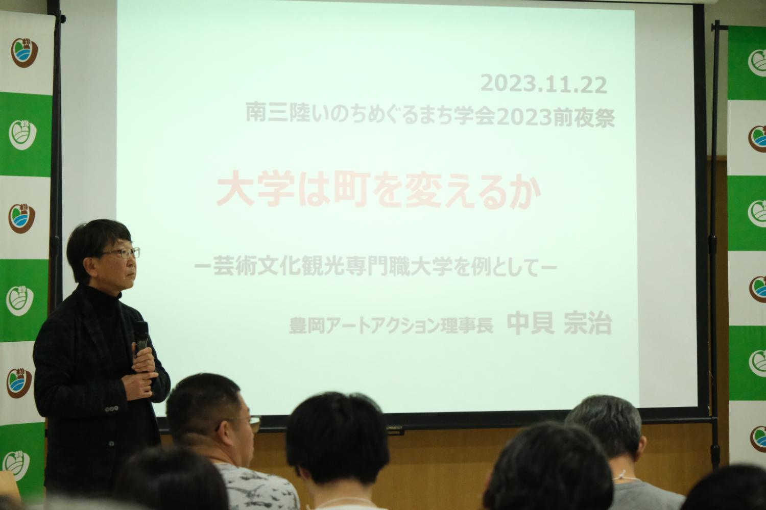 スクリーンの前で、黒い服を着た中貝宗治さんがマイクを持って話しており、多くの人々が座って聞いている様子の写真