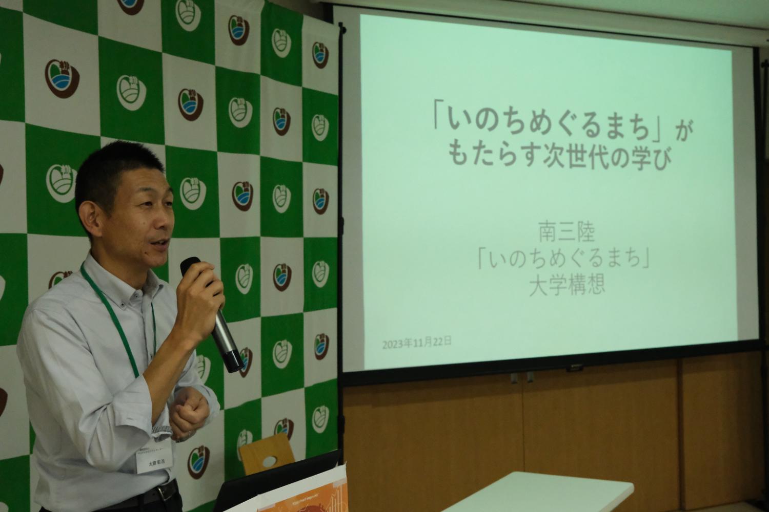 太齋彰浩さんがマイクを持って話しており、スクリーンには「『いのちめぐるまち』がもたらす次世代の学び 南三陸『いのちめぐるまち』大学構想」と書かれている写真