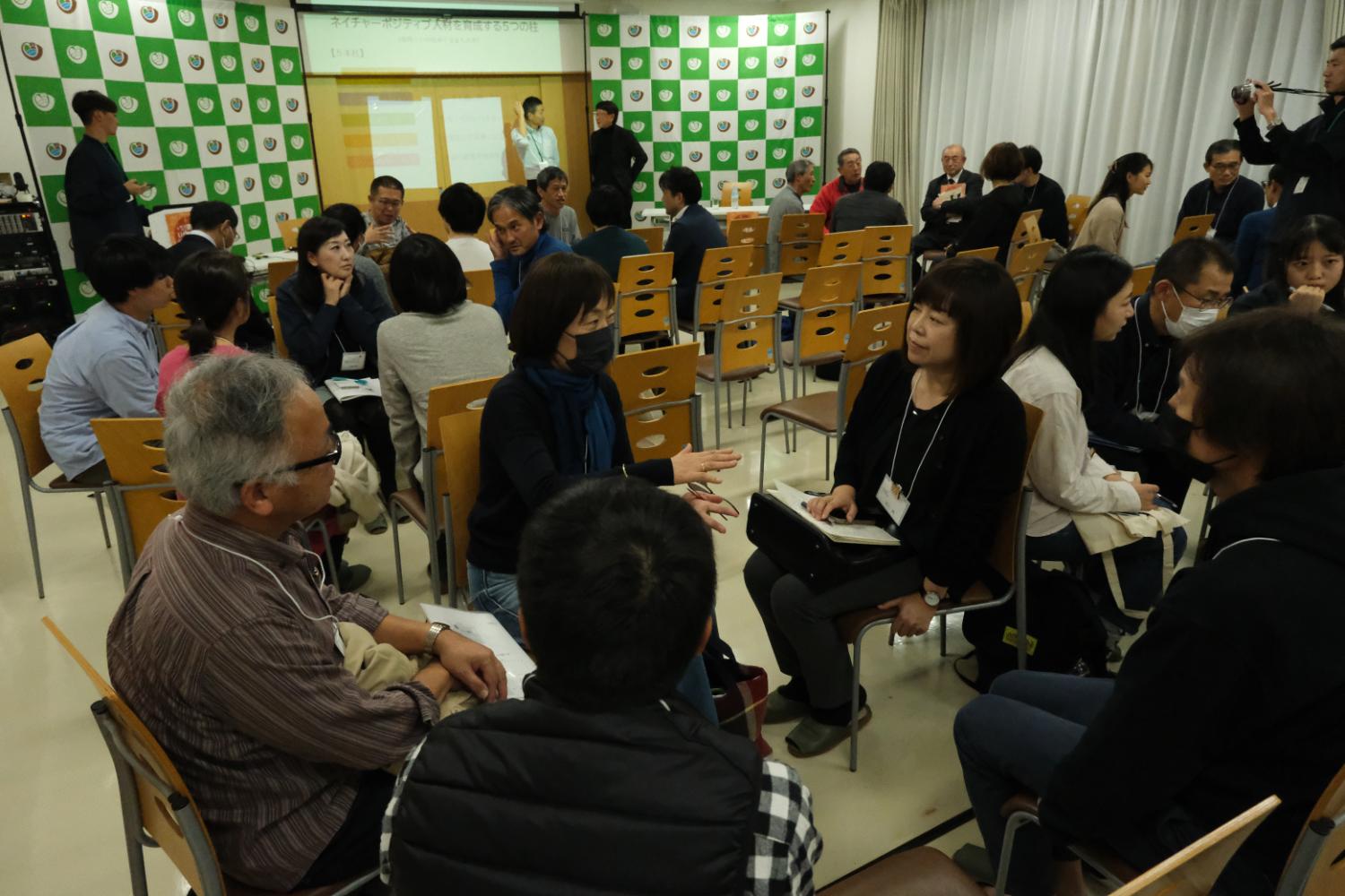 多くの人々が椅子に座り、グループで話し合っている様子で、背景には資料が映されたスクリーンが見える室内の写真