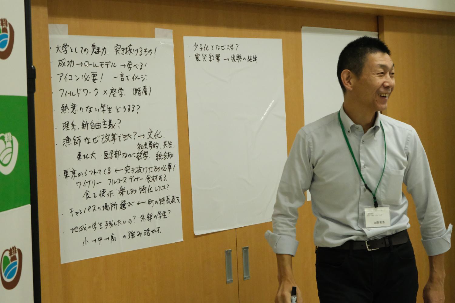太齋彰浩さんが立っている前に、木製の壁に貼られた2枚のホワイトボードがあり、学びの場を作るための意見が箇条書きに書かれている写真