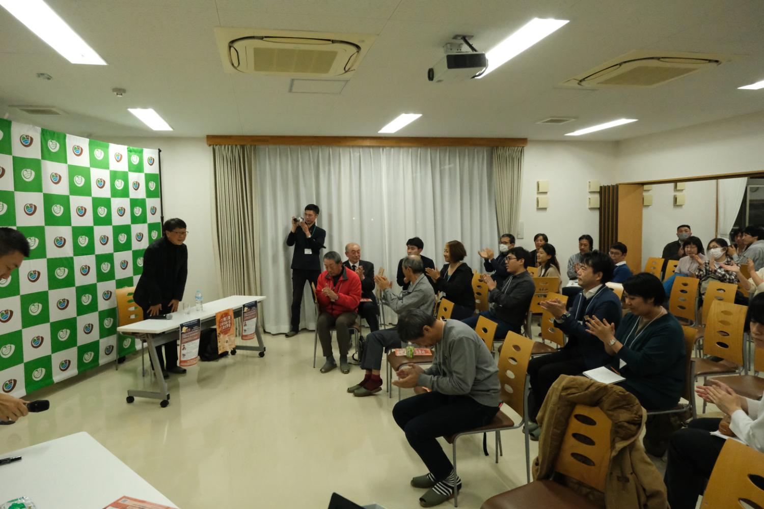 多くの人々が椅子に座って拍手をしており、緑と白の模様があるボードの前で講師が立って話している写真