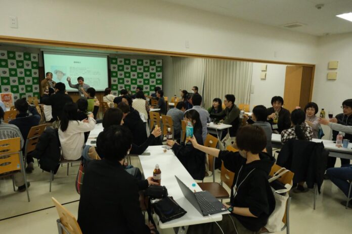 前方にスクリーンとプロジェクターが設置された室内で、参加者たちがテーブルに座って飲み物を片手に歓談しながら交流を深めている前夜祭の様子を写した写真