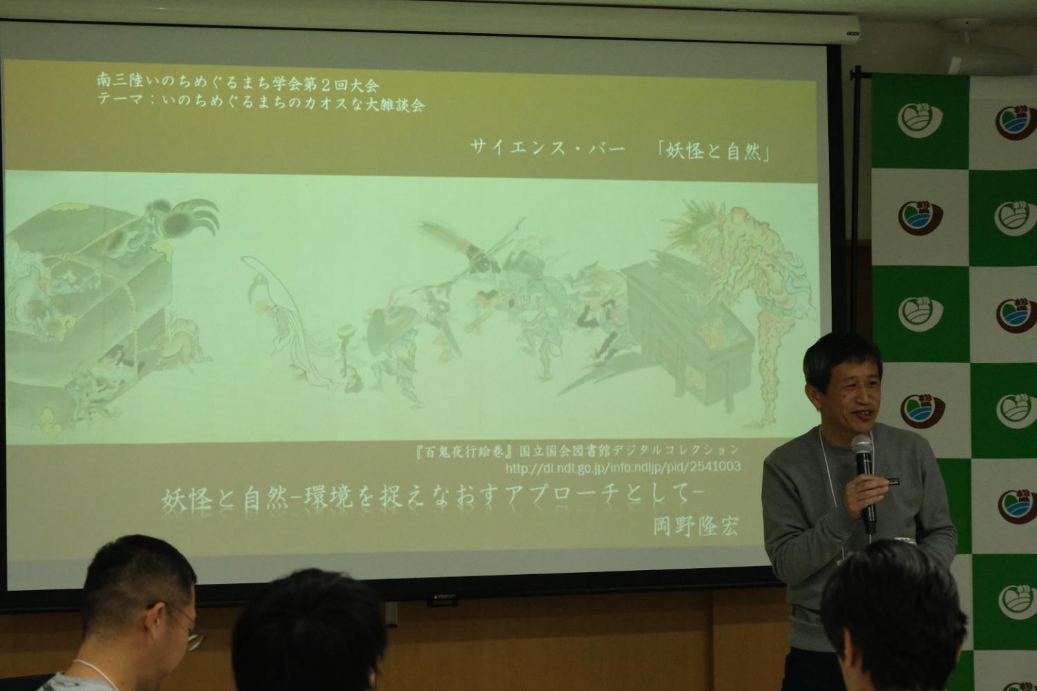 男性がマイクを持って話しており、スクリーンには サイエンス・バー『妖怪と自然』と書かれた文字の下に、妖怪のイラストが写し出されている写真