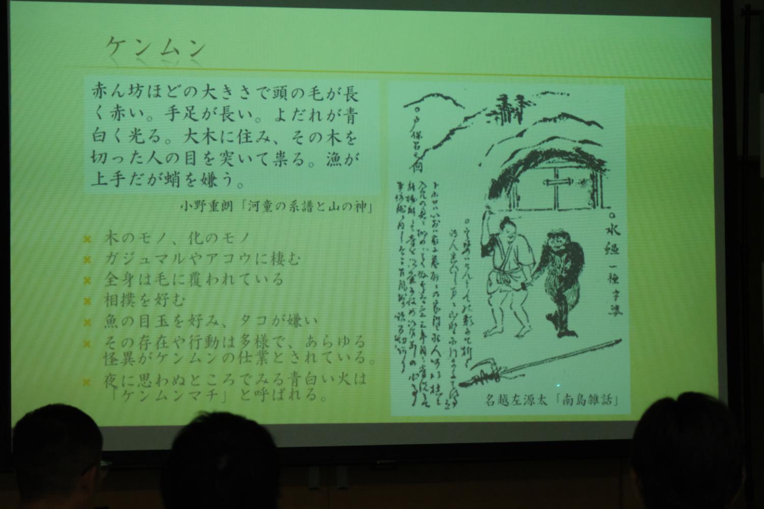 スクリーンに「ケンムン」と題された文章と赤ん坊ほどの大きさで頭の毛が長い生物のイラストが映し出されている写真