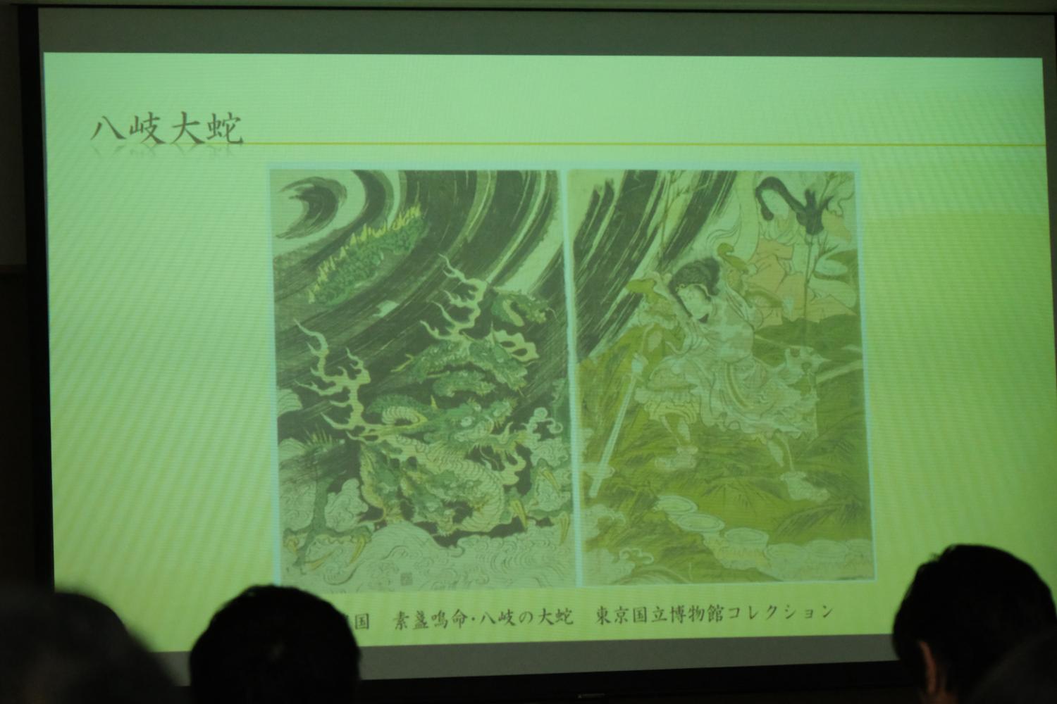 スクリーンに「八岐大蛇」と題された文章と、左側に巨大な蛇、右側に剣を持った人物が描かれた絵が映し出されている写真