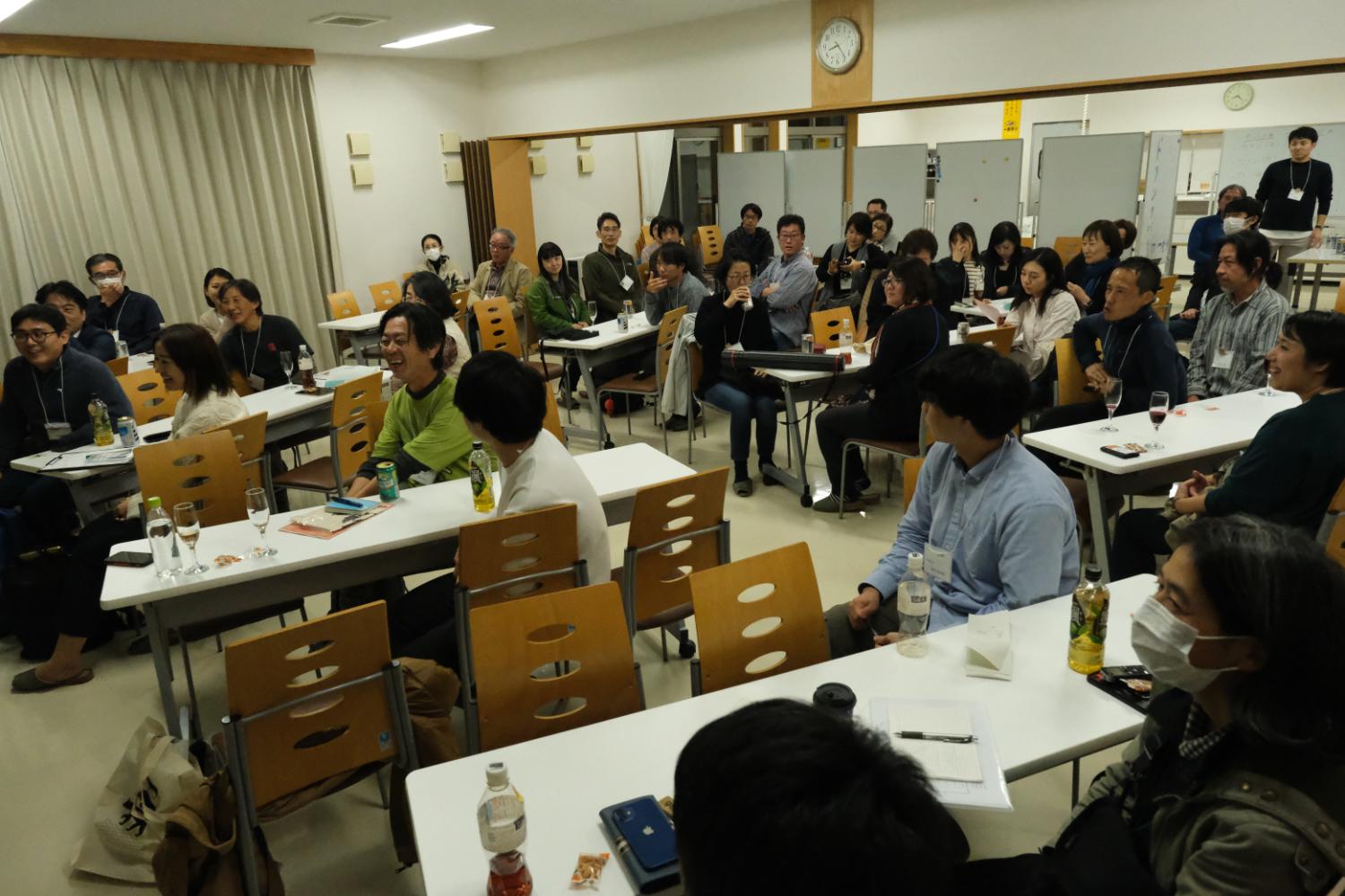 会議室で多くの参加者がグループごとにテーブルに座り、笑顔で前方を見ながら和やかに交流していて、飲み物やメモ帳が各テーブルに置かれている、懇親会中の写真