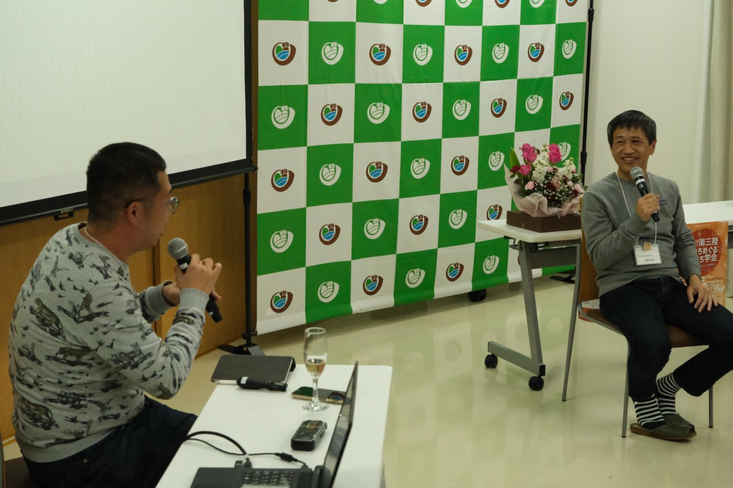 白と緑のチェック柄のロゴ入り幕を背景に、テーブル越しに向かい合って座った二人の男性がマイクを使って楽しそうに対話しているトークイベント中の写真