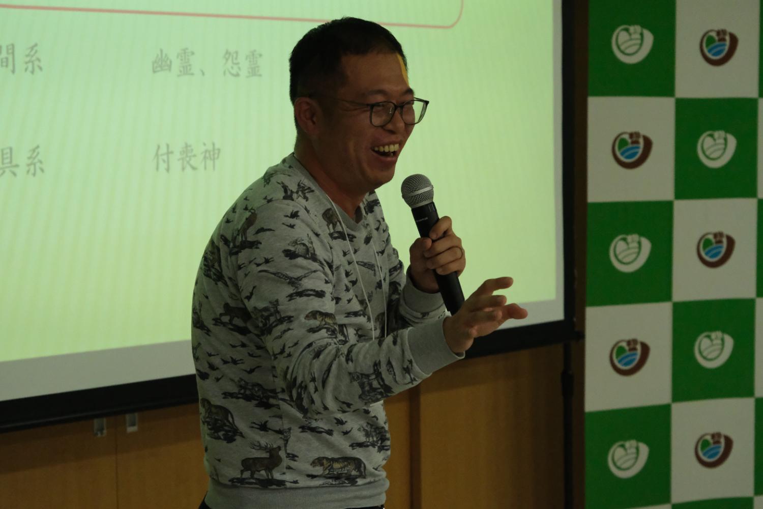 スクリーン背にしてマイクを持ちながら笑顔で話している佐藤太一さんの写真