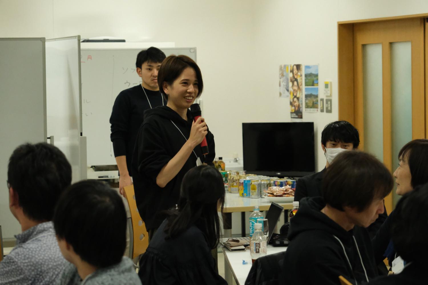 明るい室内で黒いパーカーを着た若い女性がマイクを持って笑顔で発言し、周囲には座って話を聞く人々がいる交流会の写真