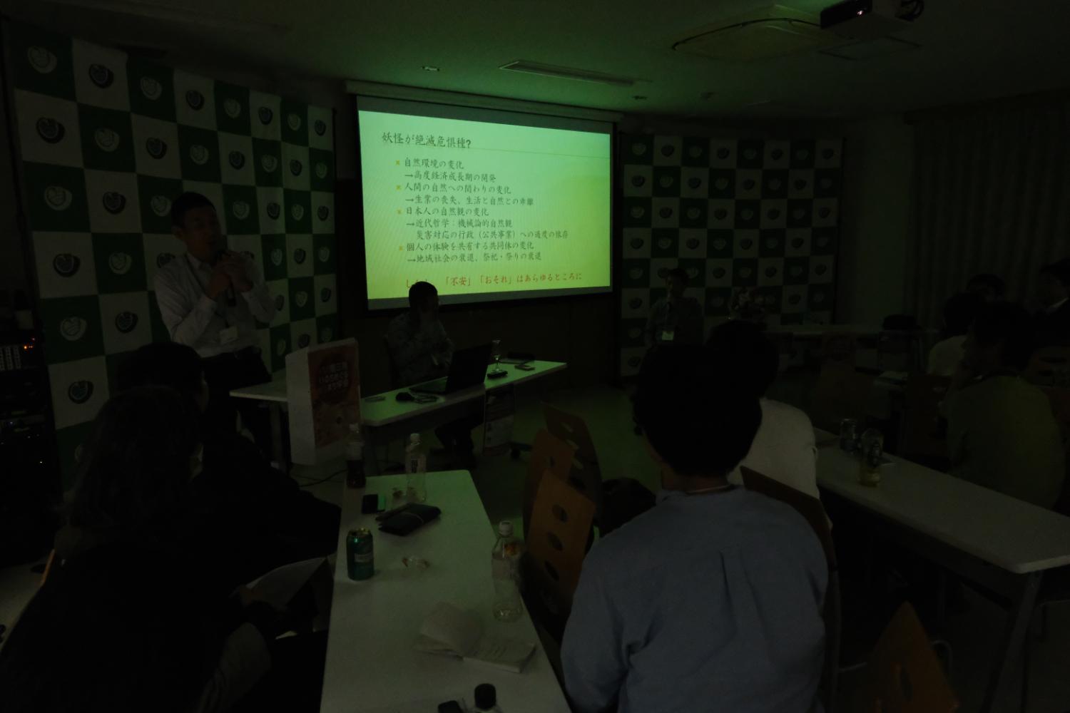 暗い会議室でプロジェクターに日本語のスライドが映し出され、数人の参加者がテーブルに座って真剣に話を聞いている、プレゼンテーション中のセミナーの写真