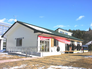 雪が部分的に残る地面の上に建つ白い壁と黒い屋根の建物の玄関前に紅白幕が飾られ、関係者の方々がが集まっている落成式の写真