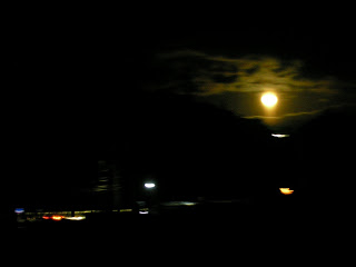 夜空に満月が明るく浮かび、雲がかかった山々が広がる中、遠くには街の灯りが点々と輝いている、夜の風景の写真