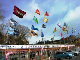建物の屋根の上に、さまざまな色とデザインのJリーグのフラッグが風になびいている様子を写した写真