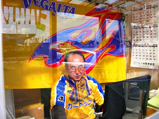 黄色に青のラインが入ったサッカーユニフォームを着て、応援グッズを身に着けた男性が、黄色と青色の大きな旗の前に座っている様子をガラス越しに撮影した写真