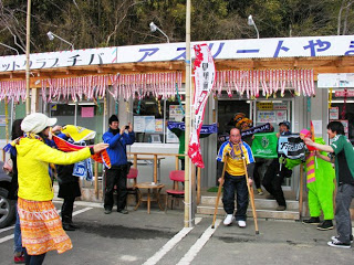 サッカーの応援グッズを持った人々が、商店街から出てきた黄色のユニフォームを着て松葉づえをついた男性に向けて歓声を上げている様子を写した写真