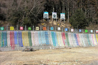 山のふもとのコンクリート製の壁に、「田束山」と書かれた大きな看板と「全世界のみなさんありがとう」と書かれたカラフルな文字板が掲げられ、壁面に色とりどりの千羽鶴が帯のように並べて吊るされている千羽鶴の掲示の写真