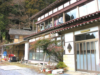 山あいの静かな場所に建つ日本家屋の前に庭が広がり、植木鉢や小さな木が並んでいるエステハウスの外観写真