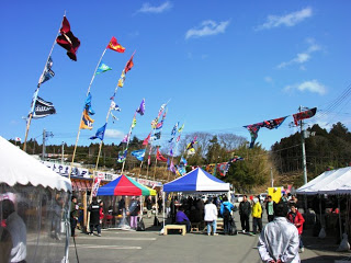 色とりどりのJリーグのフラッグが風にたなびく青空の下、出店の白いテントやカラフルなテントが並び、多くの人が行き交うにぎやかなイベント会場の写真