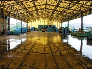 広い屋根付きの市場の中、青いプラスチック製のコンテナやパレットが一か所にまとめられて整然と並べられており、作業をしている人などがいない静かな様子を写した写真