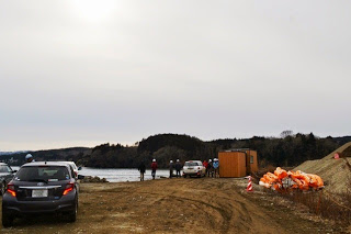 車が数台止まり、ヘルメットをかぶった人たちが海沿いの仮設道路のような場所に集まっている写真