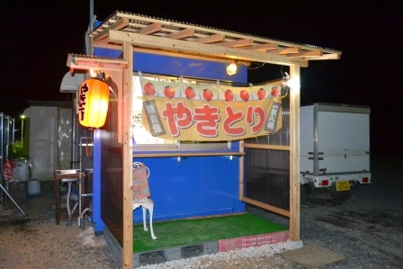 夜の屋外にある、提灯と「やきとり」の暖簾がかかっている青色の壁の小さなやきとり屋台の写真