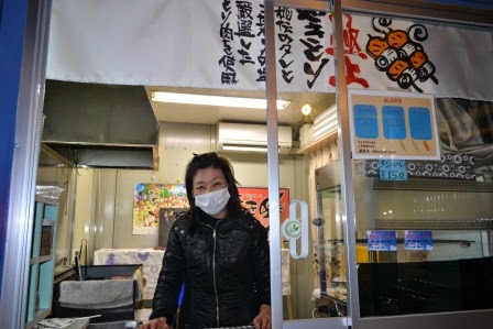 白い暖簾がかかった小さな店舗の窓から、マスクをした女性が笑顔で顔を出している写真