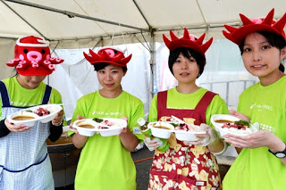 緑色のTシャツを着た4人の女性が、赤いタコの帽子やエプロンを身につけテントの下でカレーライスを手に持っている写真
