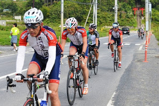 ヘルメットとサイクリングウェアを着た4人のライダーを先頭に、自転車で一般道を走行しており、その後方にも数名の自転車競技参加者が続いている写真