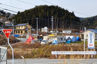 遠くには緑豊かな木々に囲まれた住宅や建物が点在し、手前の空き地横のスペースに赤い三角形の建物やプレハブの建物などが見えている様子を写した写真