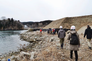川沿いの石が積まれた岸辺をヘルメットをかぶった人たちが歩いていて、奥に土手が見える写真