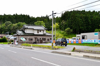 道路沿いに2階建ての住宅が建ち、その横の空き地に停まった黒い車と、歩道側に看板が設置された、遠くに緑豊かな山々が見える郊外の風景写真