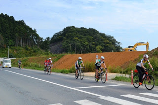 緩やかな坂道の一般道を6人の自転車競技参加者が自転車で走行しており、背景には造成中の茶色い土盛りやショベルカー、森に囲まれた山が見える写真