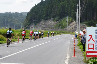 まっすぐ伸びた道路の左側を10人ほどの自転車競技参加者が連なって走行しており、右側の歩道には「入口」と書かれた看板や旗を振って応援する地元の人々がいる大会沿道の応援風景の写真