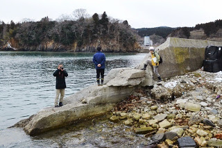壊れた護岸に立つ作業員と写真を撮る人がいて、背後に木々の茂った崖が見える写真