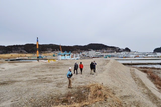枯草のある砂利が広がる場所にヘルメット姿の人たちが立ち、背景に住宅街が広がっている写真