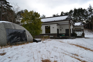 木々に囲まれた、瓦屋根に雪が積もった平屋のシルク館の外観写真