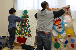 男の子と男性が、フェルトでできた大きなクリスマスツリーとキャラクターの壁飾りを取り付けている写真