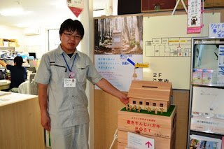 「書き損じはがき回収ポスト」と書かれた木製の箱に乗った芝生風の台座の上に、小さな人形が飾られた、木で作った屋根付きの小さな家の形のポストが設置されており、作業着を着た男性が横に立っている写真