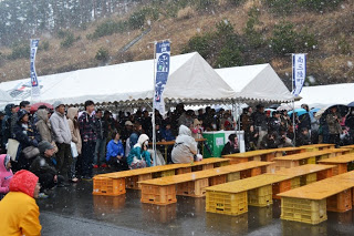 雪が降る屋外で、テントの下に多くの人が集まり、前方には木の板とコンテナを組み合わせた簡易ベンチが並べられているイベントの様子の写真