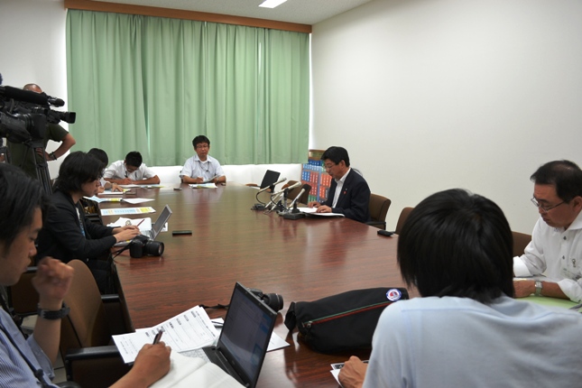 佐藤町長がマイクの前で発言し、周囲には記者たちがパソコンやノートを使って話を聞いている定例記者会見の様子の写真