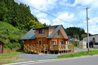 緑の山を背にした静かな道路沿いに、ウッドデッキ付きの温かみある木造の一軒家が建っている建物外観の写真