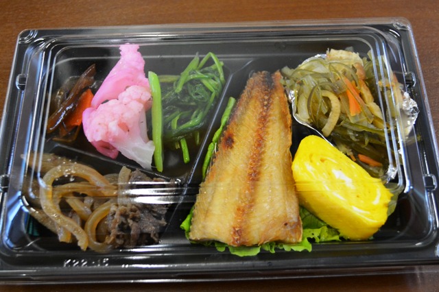 プラスチック容器に、大きなホッケ、玉子焼き、野菜のおひたし、煮物、漬物などが彩りよく詰められた和風の配達弁当が写っている写真