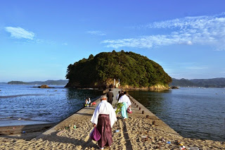 青空の下、参拝者や宮司の方々が砂の道を歩いて小島へ向かっている姿が見える、海に続く参道の写真。